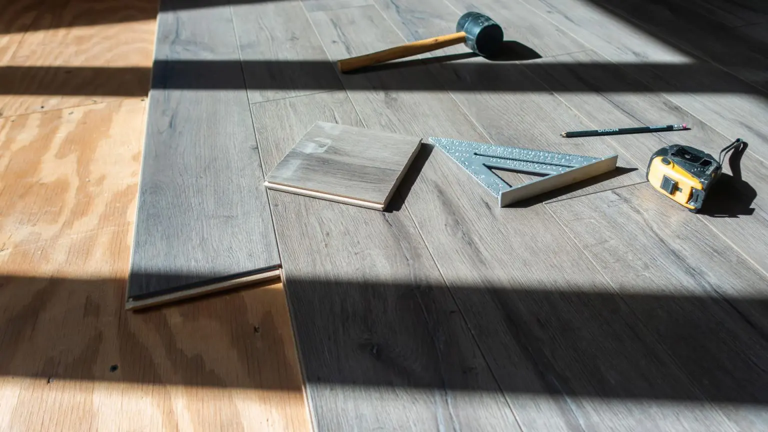 a high-angle shot of gray wood-grain flooring planks being installed over a plywood subfloor, with installation tools including a rubber mallet, speed square, tape measure, and pencil scattered on the surface