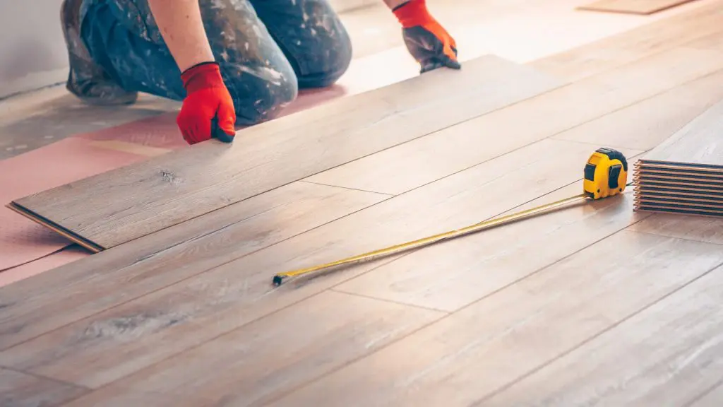 a flooring installer wearing red gloves kneeling on an underlayment to precisely fit a wood plank into place, with a tape measure resting nearby