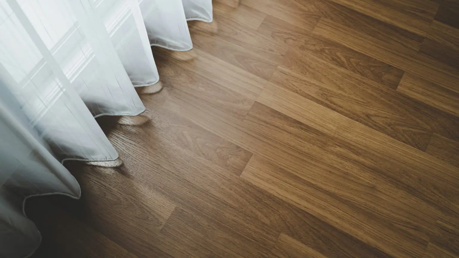 A high-angle close-up shot showing the rich grain patterns of newly installed hardwood plank flooring running alongside a sheer white window curtain