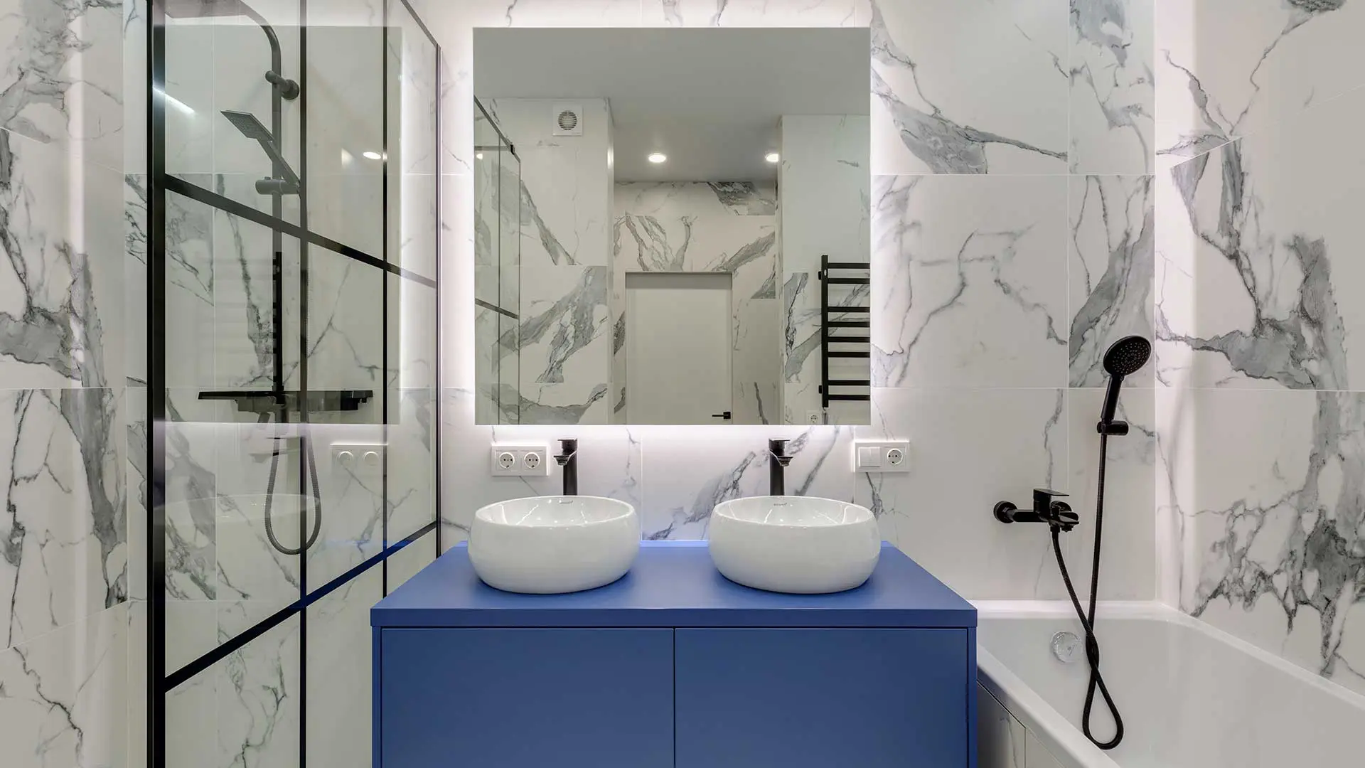 modern bathroom featuring a vibrant blue double vanity with two round white vessel sinks, set against white marble-veined wall tiles, a backlit mirror, a glass-enclosed shower, and a bathtub with matte black hardware