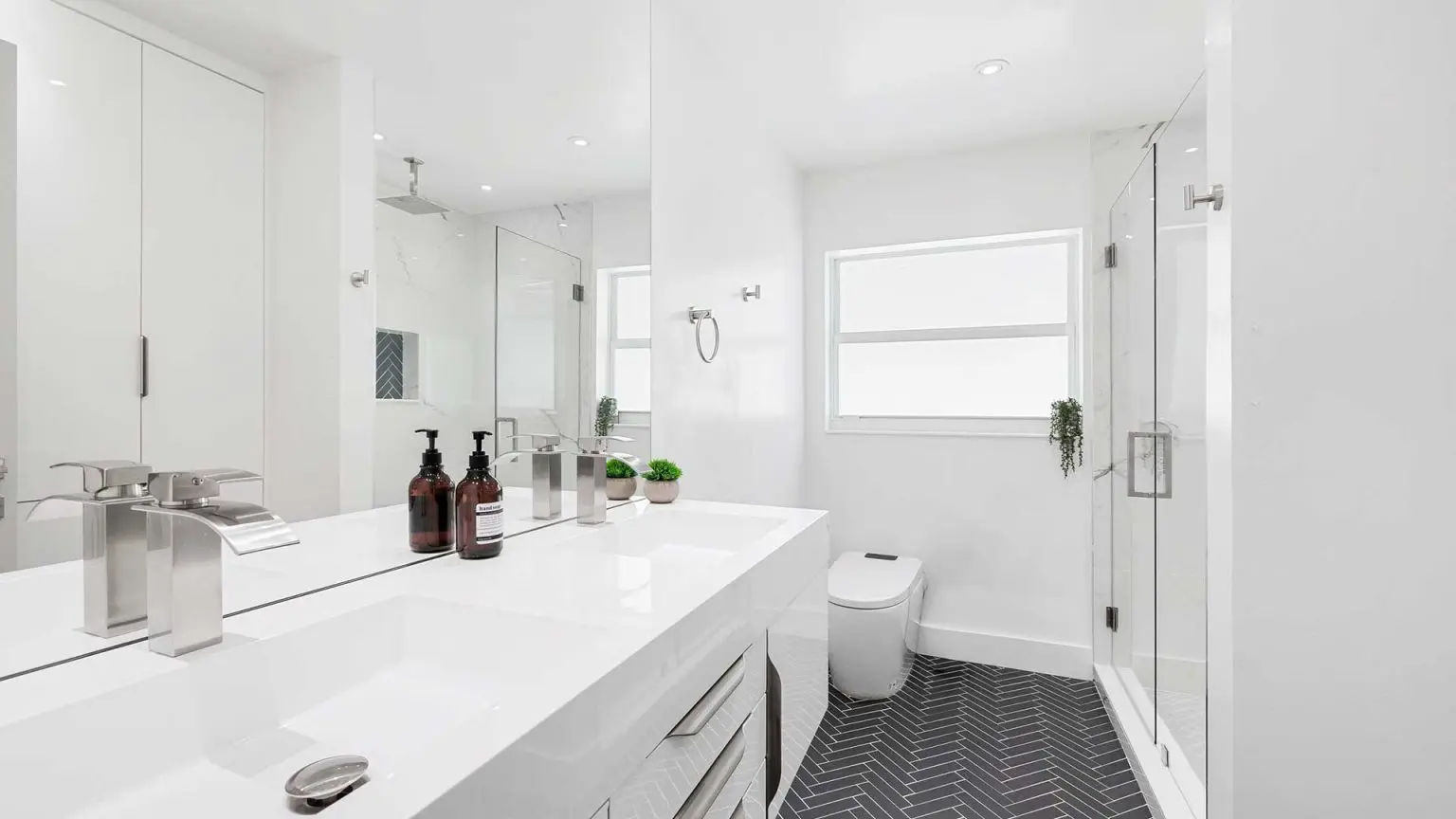 modern all-white bathroom with a long double vanity, chrome faucets, a large mirror, a glass-enclosed shower, a bidet toilet, and dark grey herringbone tile flooring
