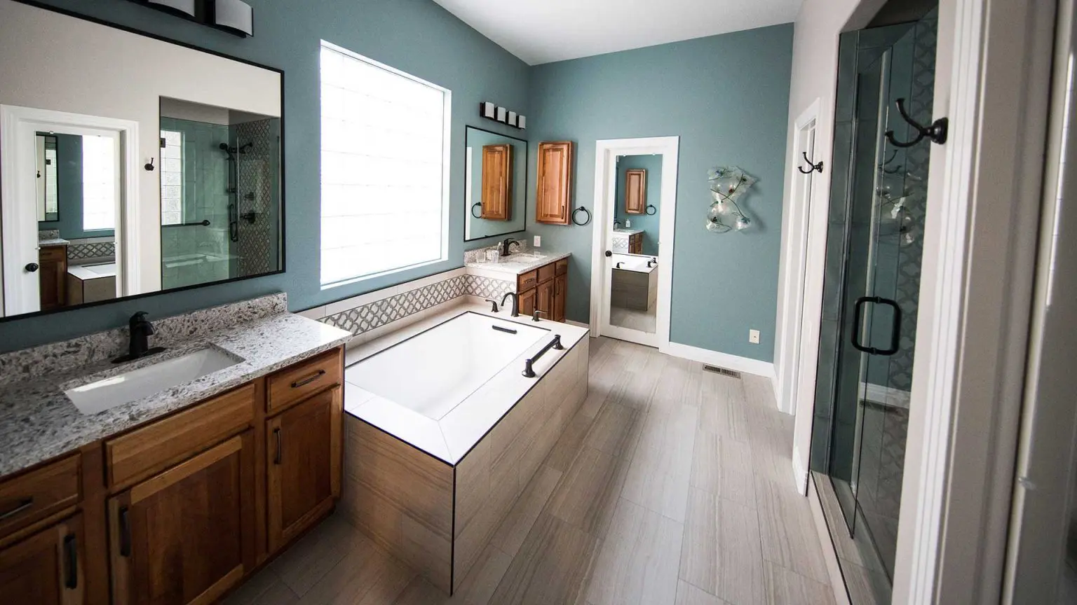 modern master bathroom with teal walls, dual wood vanities, a central paneled soaking tub, a glass walk-in shower, and light tile flooring.