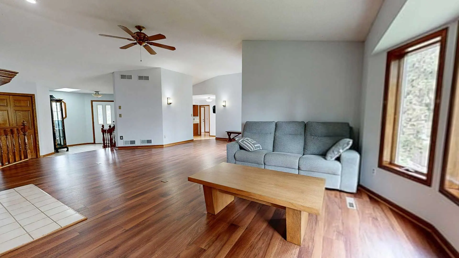 spacious, bright living room featuring medium-toned reddish-brown hardwood flooring, a gray sofa, a low wooden coffee table, and an overhead ceiling fan