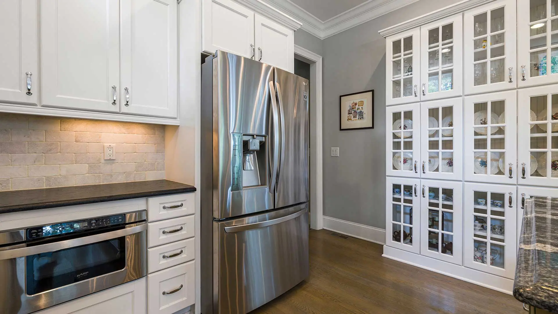 bright transitional kitchen with white cabinets, stainless steel french-door fridge, built-in microwave drawer, and a large glass-front china cabinet. features dark countertops, a light brick backsplash, and hardwood floors.