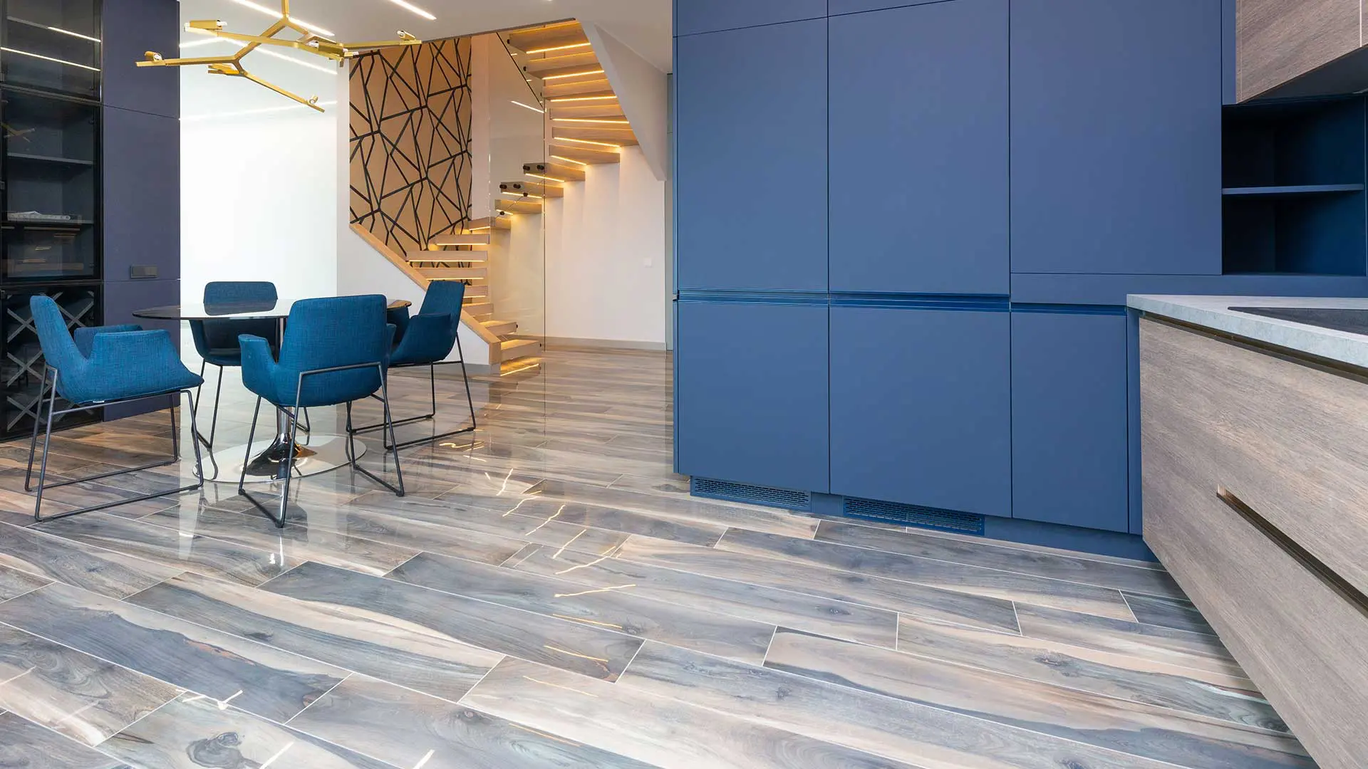 contemporary kitchen and dining space highlighted by bold blue cabinetry, a unique modern staircase, and glossy, multi-toned gray wood-grain floor tiles