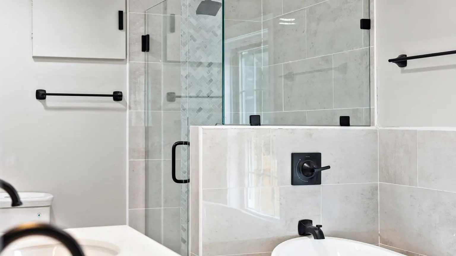 modern bathroom featuring a glass-enclosed walk-in shower with grey tiles and a herringbone accent wall, sitting adjacent to a white bathtub with matte black fixtures and matching towel bars