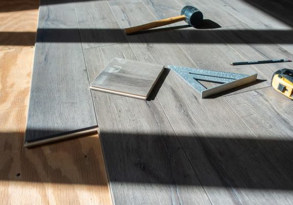 a high-angle shot of gray wood-grain flooring planks being installed over a plywood subfloor, with installation tools including a rubber mallet, speed square, tape measure, and pencil scattered on the surface