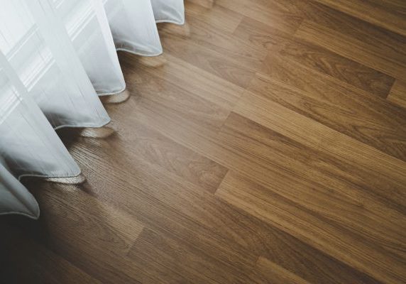 A high-angle close-up shot showing the rich grain patterns of newly installed hardwood plank flooring running alongside a sheer white window curtain